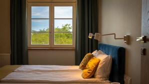 A neatly made bed with a white comforter and two yellow pillows, a bedside table with a white mug, and a window with a view of greenery and a clear sky. - Villa Bałtycka (Gdansk)