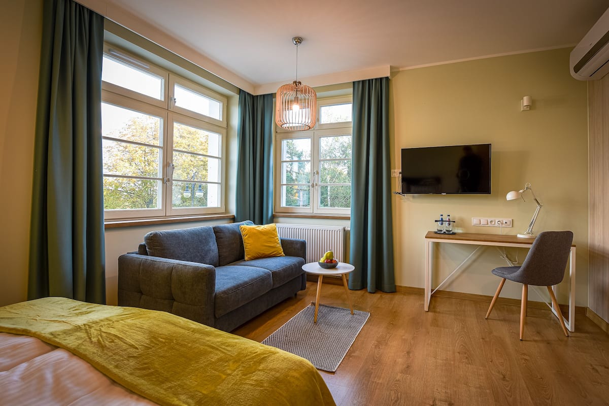 Ein Hotelzimmer mit Bett, einer Couch, einem Schreibtisch mit Stuhl, einem Fernseher und großen Fenstern mit Vorhängen.