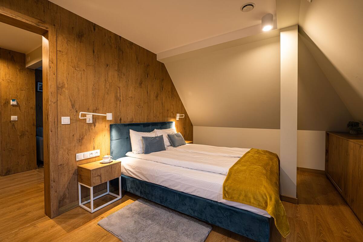 Ein Schlafzimmer mit einer Wand in Holzoptik, einem Bett mit weißer Bettwäsche und einem blauen Kopfteil, einem Nachttisch mit Lampe und einem Holzschrank.