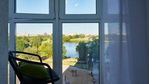 A room with a view of a lake and park, a chair with a pillow, and a window with blinds. - PaulMarie Apartments on Mayakovskogo 35 (Minsk)