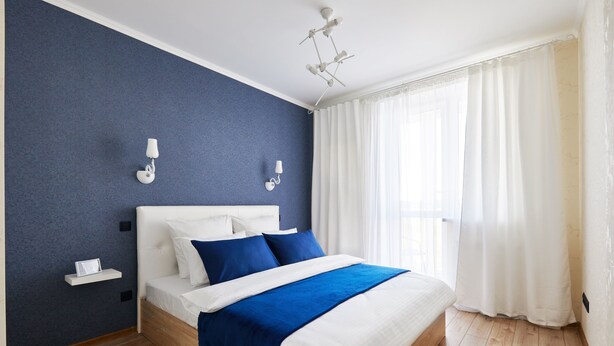 A modern bedroom with a wooden bed, white headboard, blue accent pillows, and a bedside table with a lamp. - PaulMarie Apartments on Mayakovskogo 35 (Minsk)