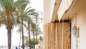 A balcony with a tiled floor, potted plants, a bamboo screen, and a street view with palm trees and parked cars. - Appartement Palmeras Puerto Paraiso Estepona (Estepona)