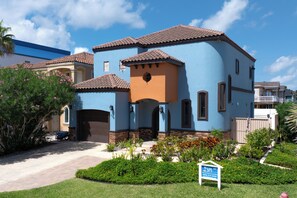 Ein zweistöckiges Haus mit blau-orangenem Fassadengestalt, einem Ziegeldach und einem Vorgarten mit einem Schild, das ein zum Verkauf stehendes Objekt anzeigt.