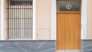 A building facade with a barred window, a wooden door with a number plate, and a tiled base.