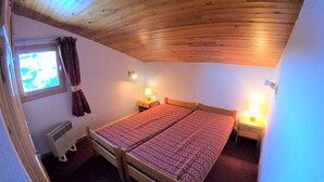 Une chambre avec un plafond en bois, un lit double avec une literie à motifs, deux lampes de chevet, une fenêtre avec des rideaux et un radiateur.
