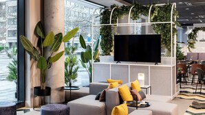 A modern living area with a large flat-screen TV, comfortable seating, and a view of the city through large windows. - NH Hannover (Hannover)