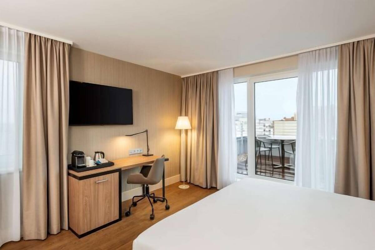 A hotel room with a bed, desk, chair, TV, lamp, and a balcony with a table and chairs.