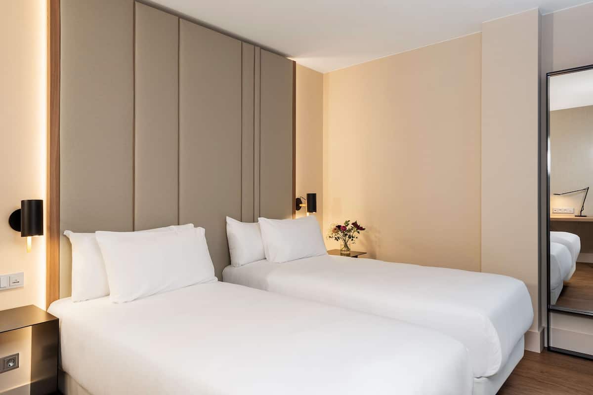 A hotel room with two beds, a bedside table, a wall-mounted light, and a small table with a vase of flowers.