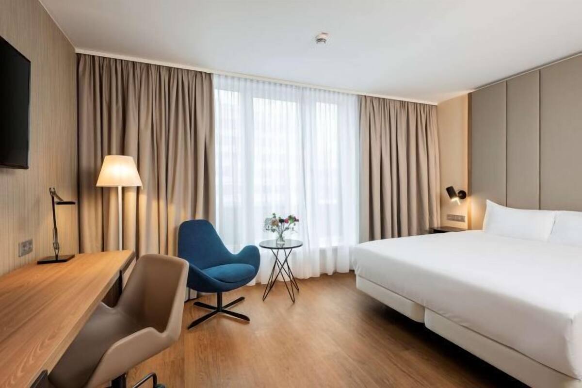 A hotel room with a bed, a desk, a chair, a small table with a vase, and curtains.