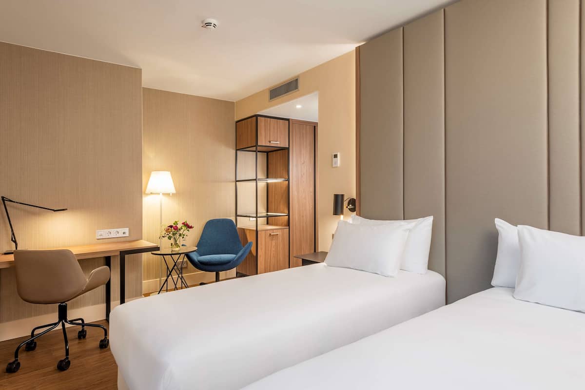 A hotel room with a bed, a desk, a chair, and a lamp.