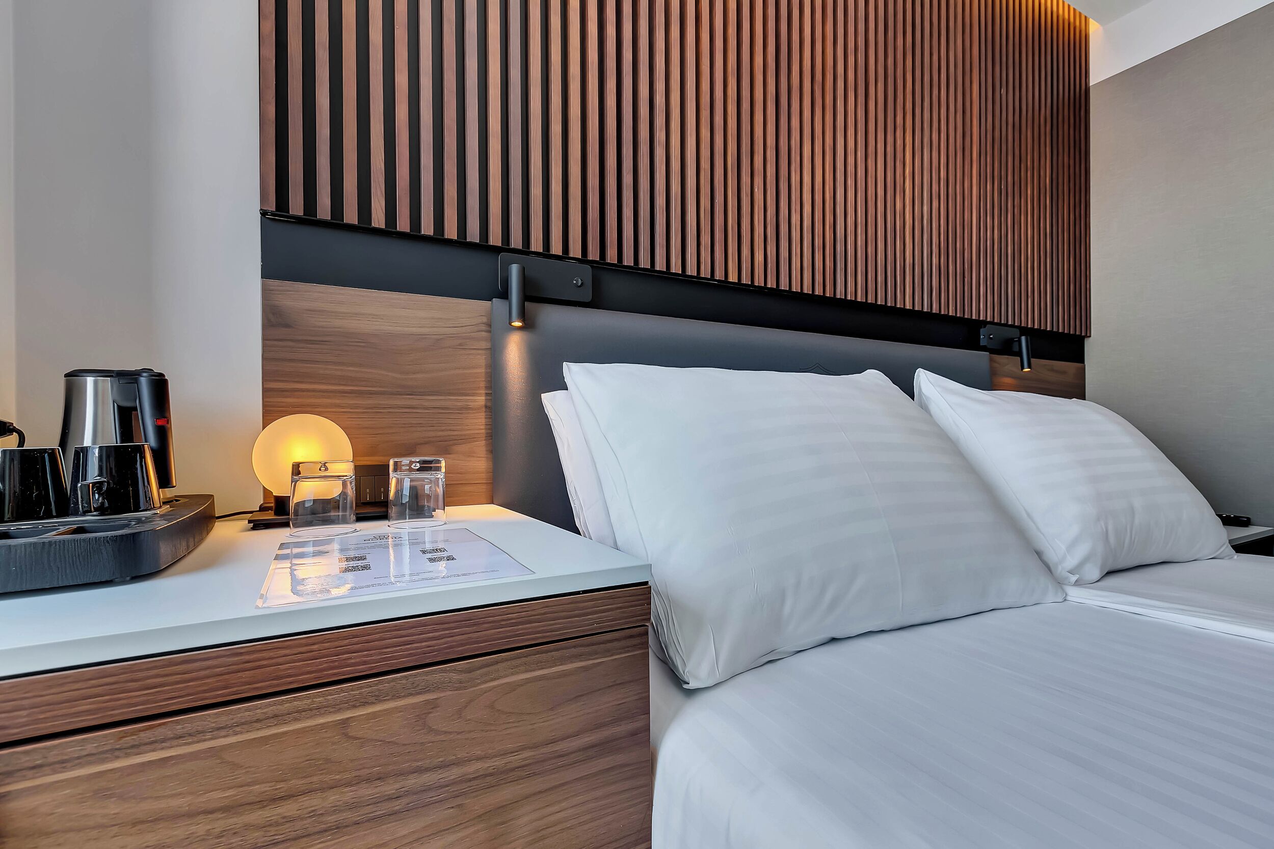 Ein modernes Hotelzimmer mit einem Holzkopfteil, einem Nachttisch mit Kaffeemaschine, Gläsern und einer Speisekarte.