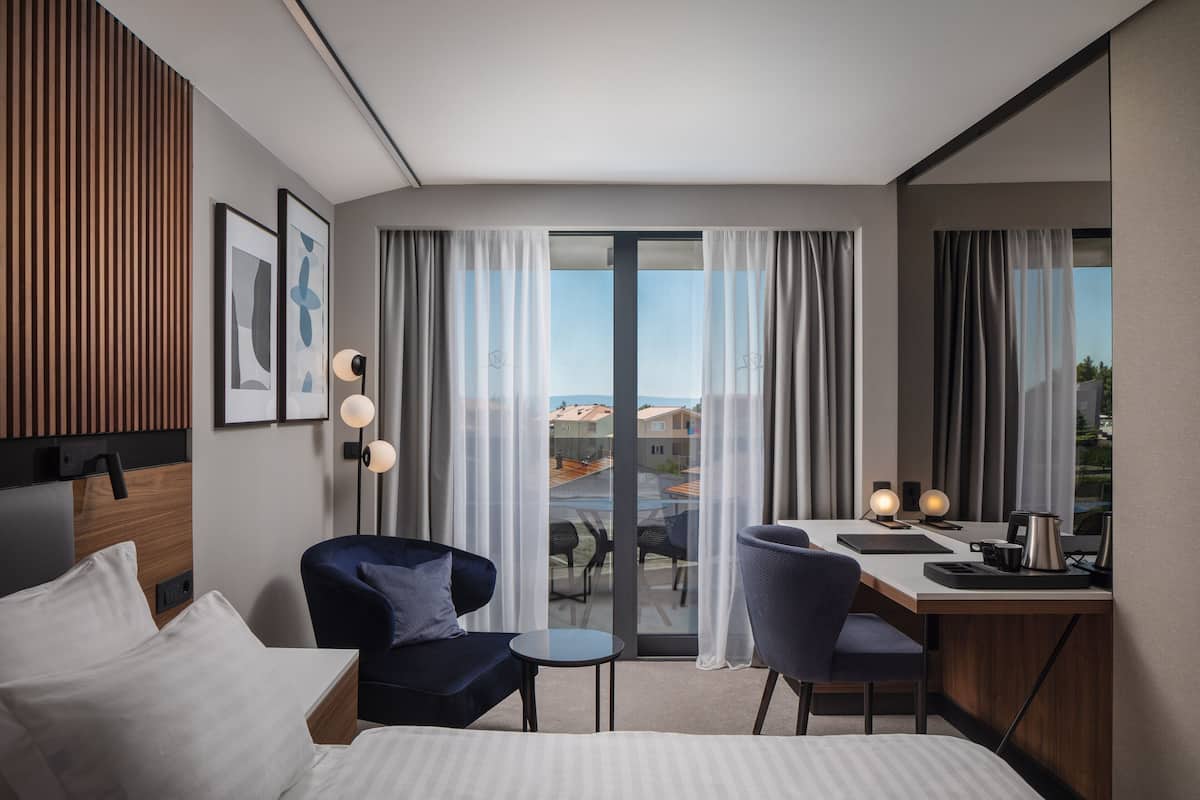 A modern hotel room with a bed, a desk, a chair, and a balcony with a view.