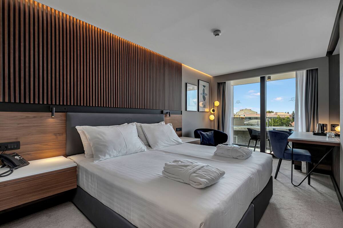 A modern hotel room with a large bed, a desk, and a view of the city through large windows.