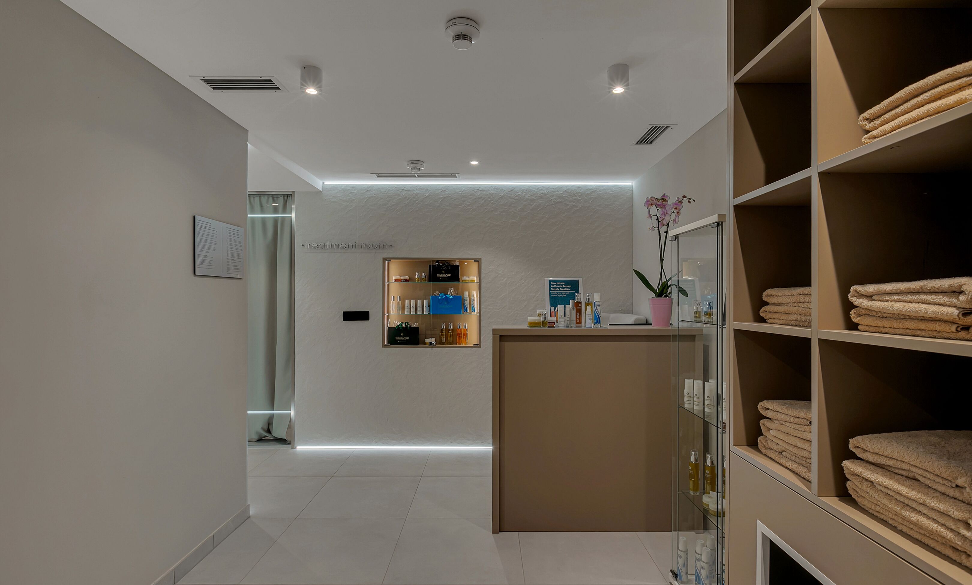 A modern treatment room with a reception desk, shelves with towels, and a glass partition.