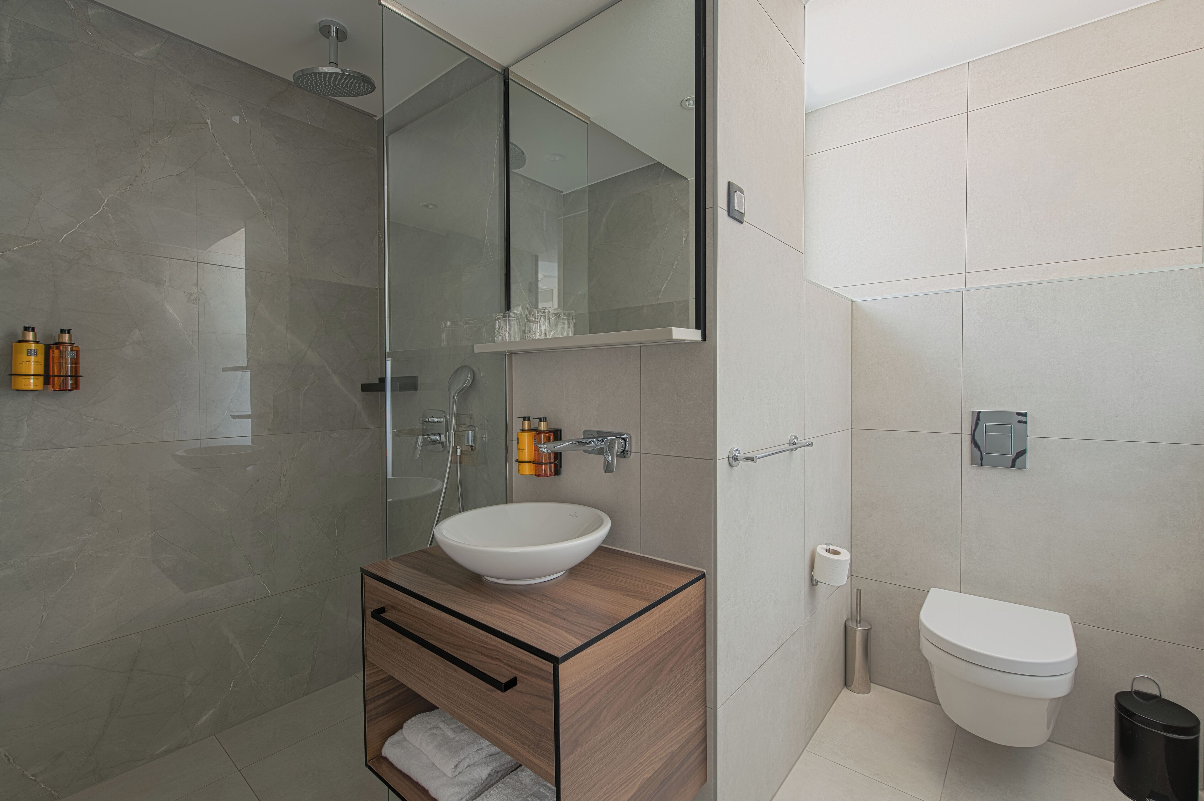 Un bany modern amb dutxa de vidre, un tocador de fusta i un lavabo blanc, i un vàter.