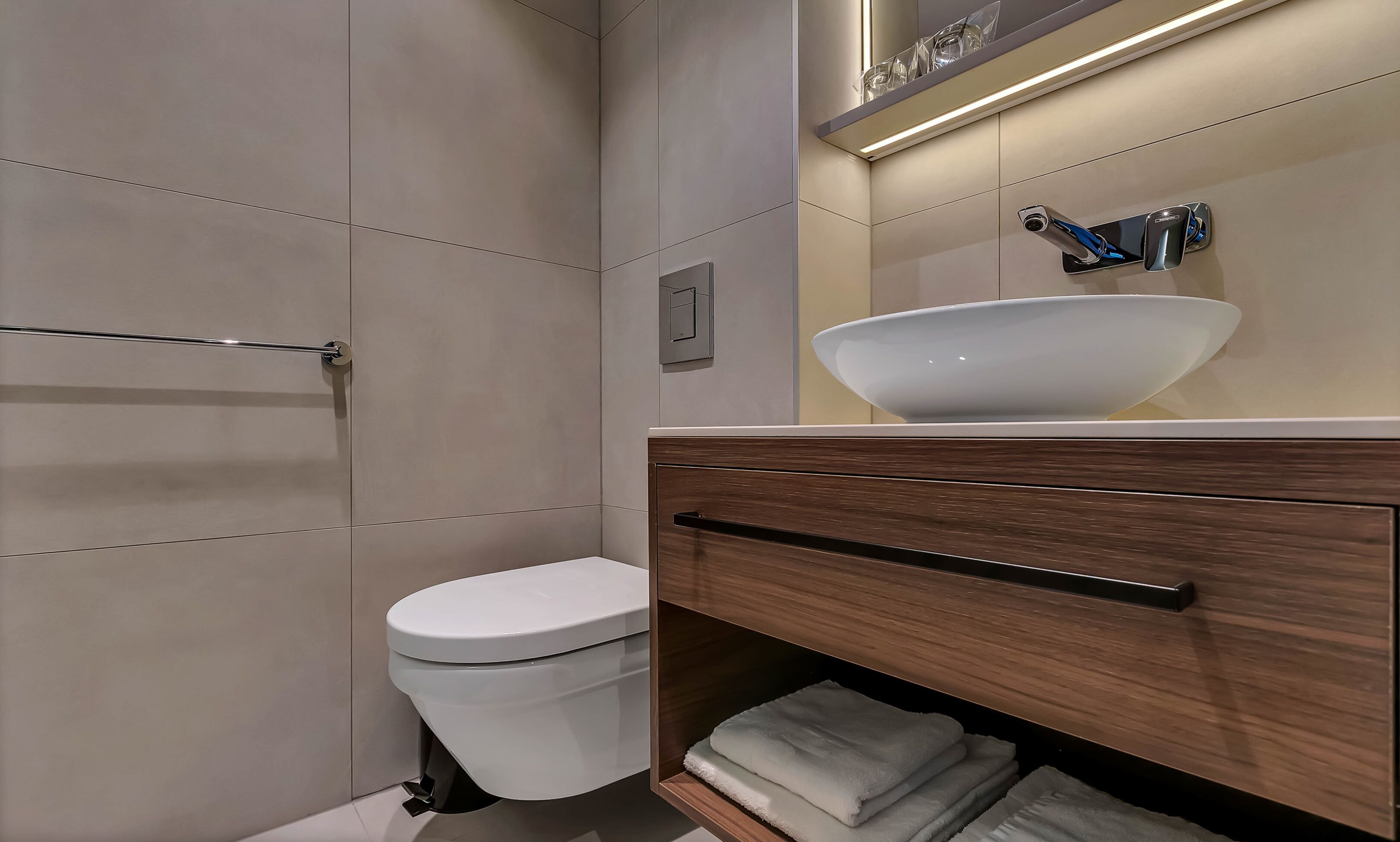 Un baño moderno con inodoro blanco, tocador de madera con un espejo grande y lavamanos blanco.
