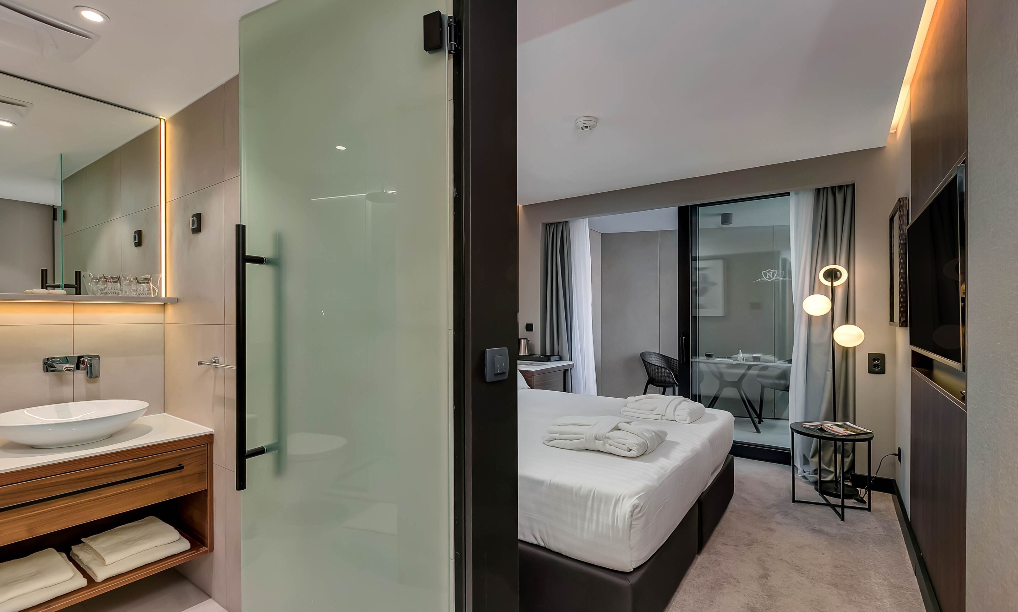 A modern hotel room with a glass shower enclosure, a large bed with a neatly made white towel, a small bedside table, and a large mirror.