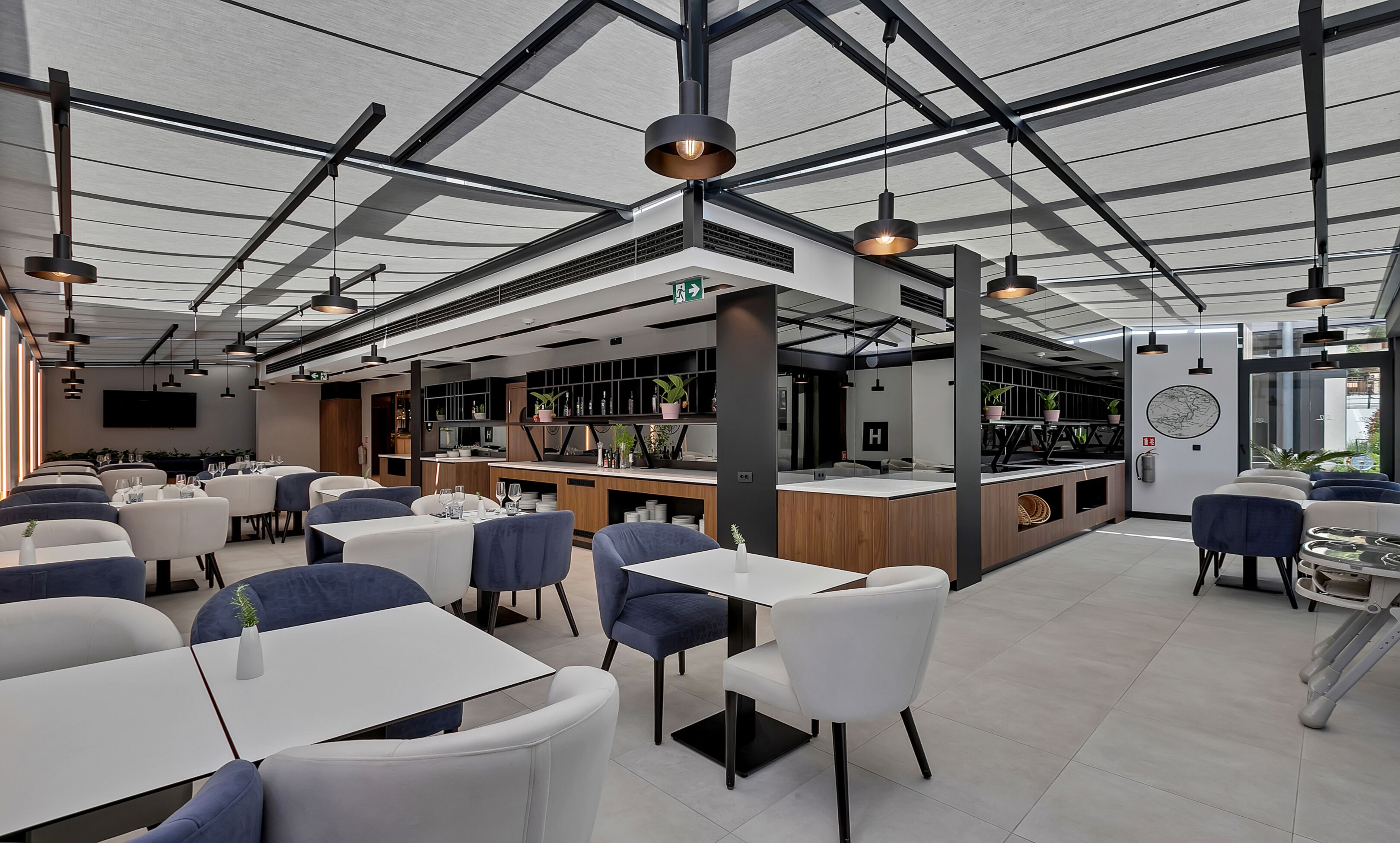 A modern restaurant with a high ceiling, exposed beams, and pendant lighting. Tables are arranged in the foreground, and a bar area is visible in the background.