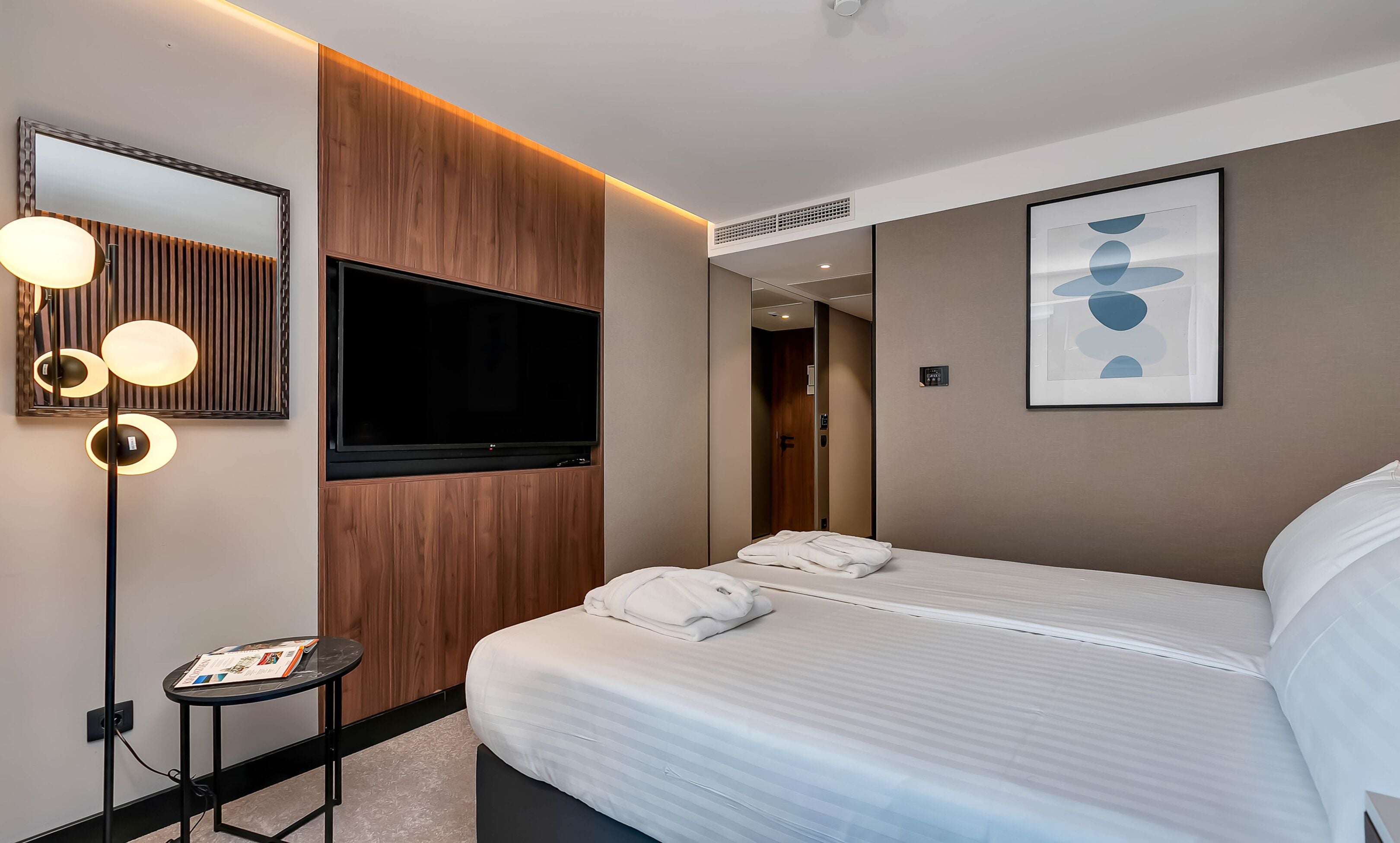 Quarto de hotel com uma cama grande, televisão de tela plana, mesa de cabeceira com uma revista e espelho fixado na parede.