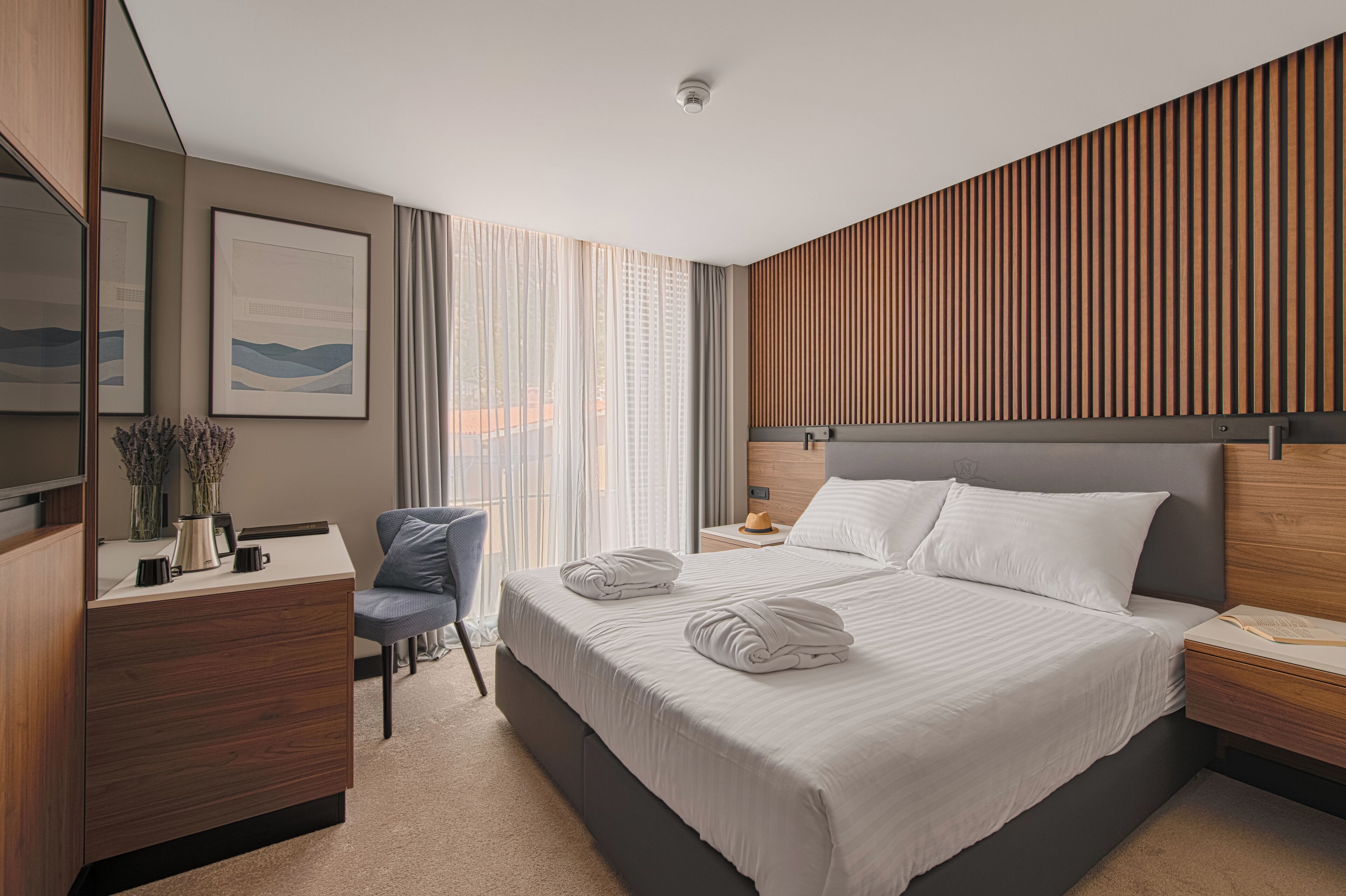 A modern hotel room with a large bed, a desk with a chair, and a view of the outdoors through large windows.