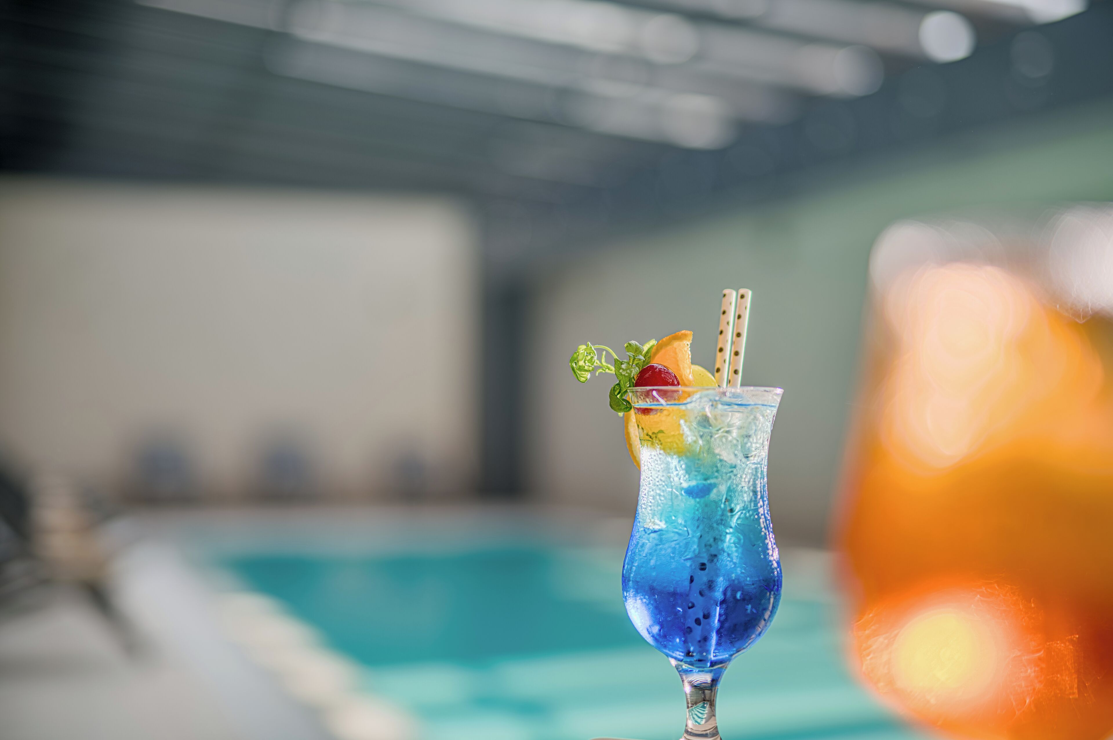 A cocktail with a blue hue, garnished with fruit and a mint leaf, placed on a table near a pool.