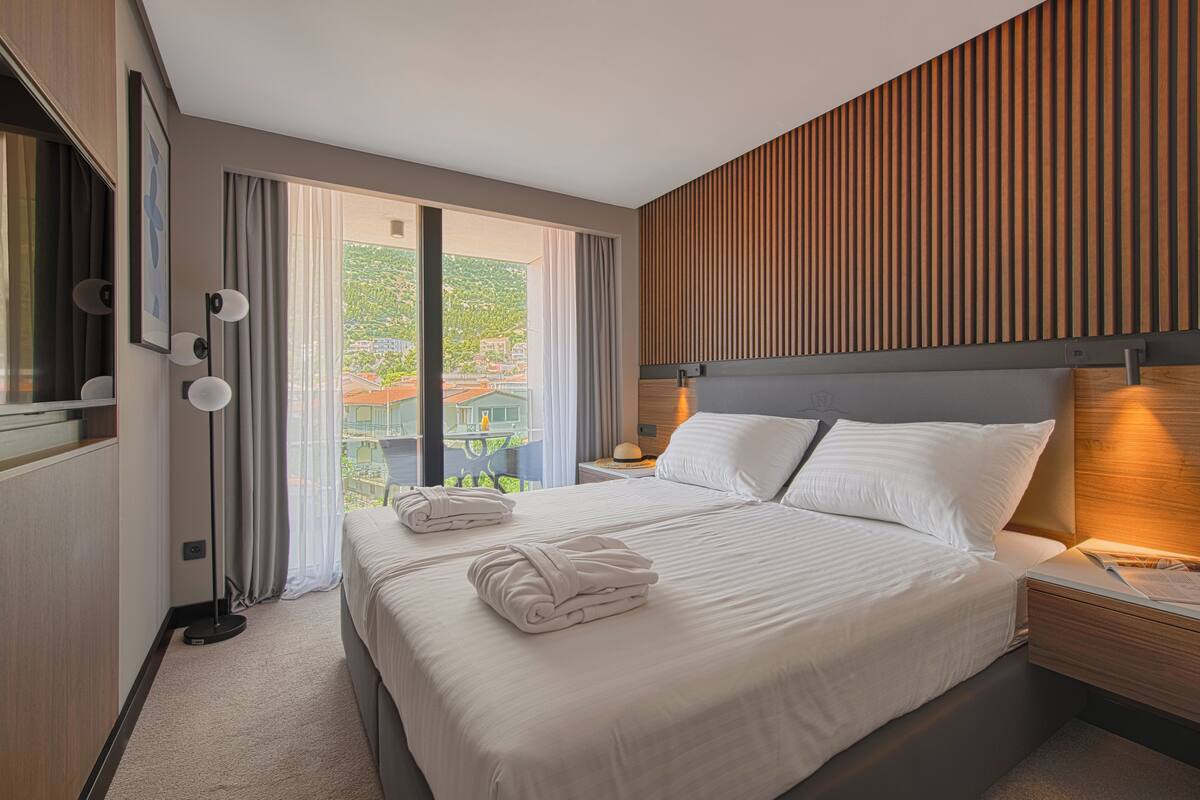 A modern hotel room with a large bed, a nightstand, a bedside lamp, and a view of a green landscape through the window.