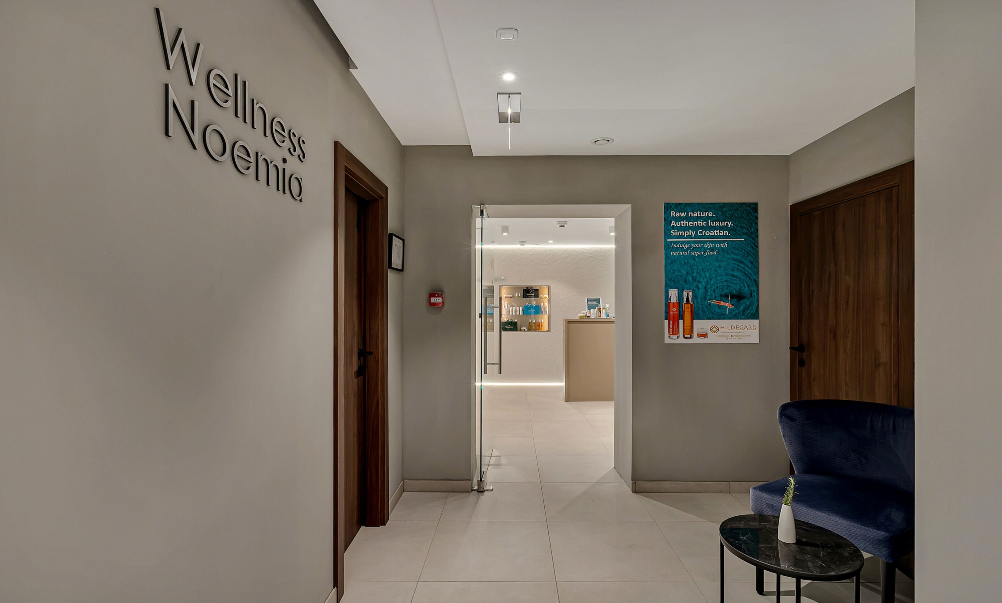 A hallway with a sign reading "Wellness Noemi" and a poster advertising "Hildegard" products. A blue chair and a small table are visible.