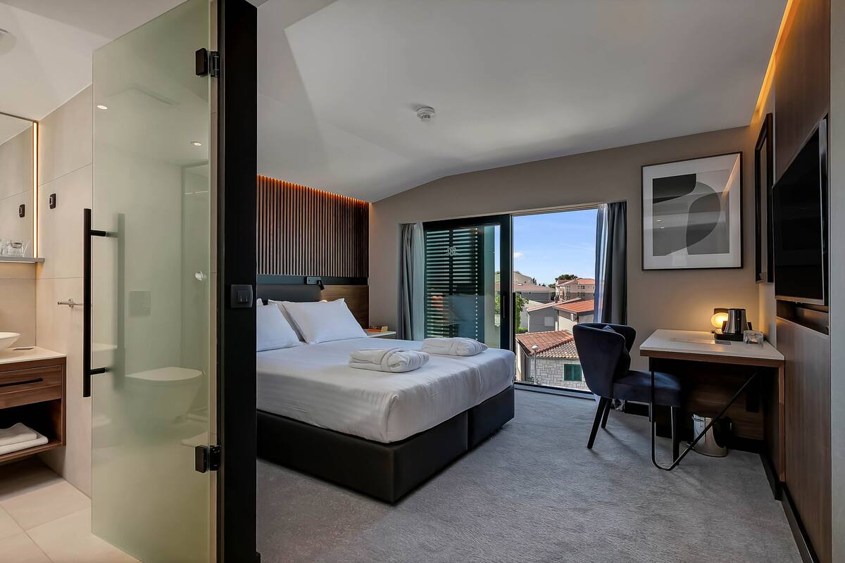 A modern hotel room with a bed, a desk, a chair, a bathroom with a sink and mirror, and a view of buildings through the window.