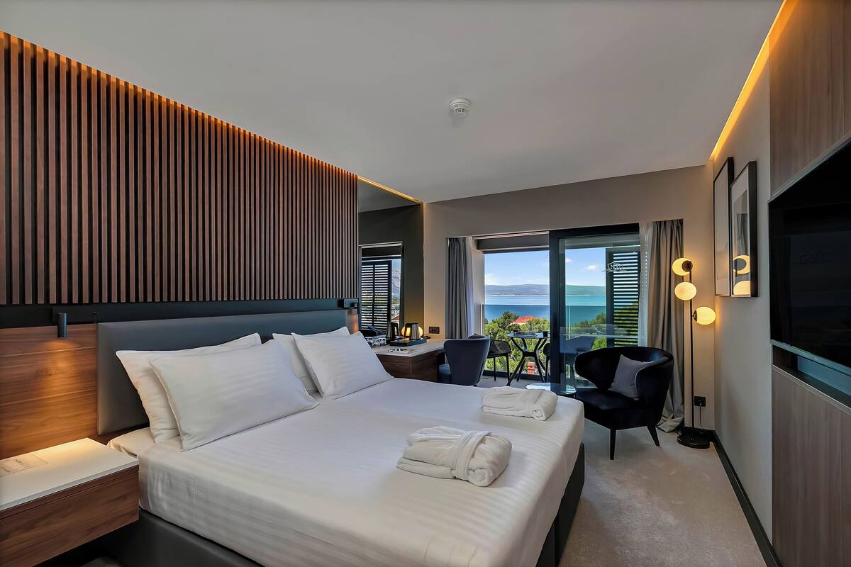 A modern hotel room with a large bed, a desk, a chair, and a view of the sea.