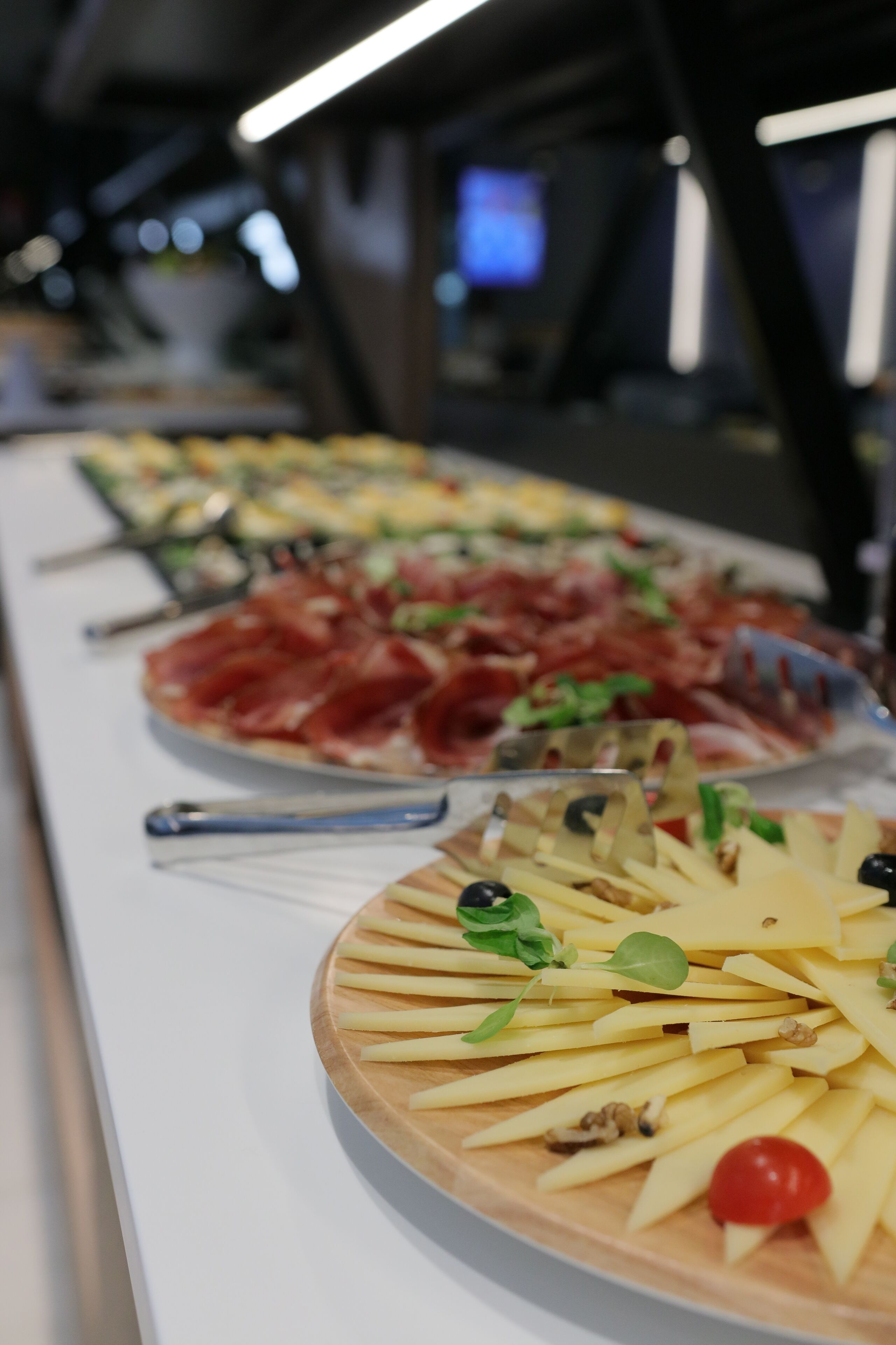 Una mesa de buffet con diversos alimentos, entre ellos una pizza con jamón serrano y hojas verdes, un plato con queso, aceitunas y un tomate cherry.