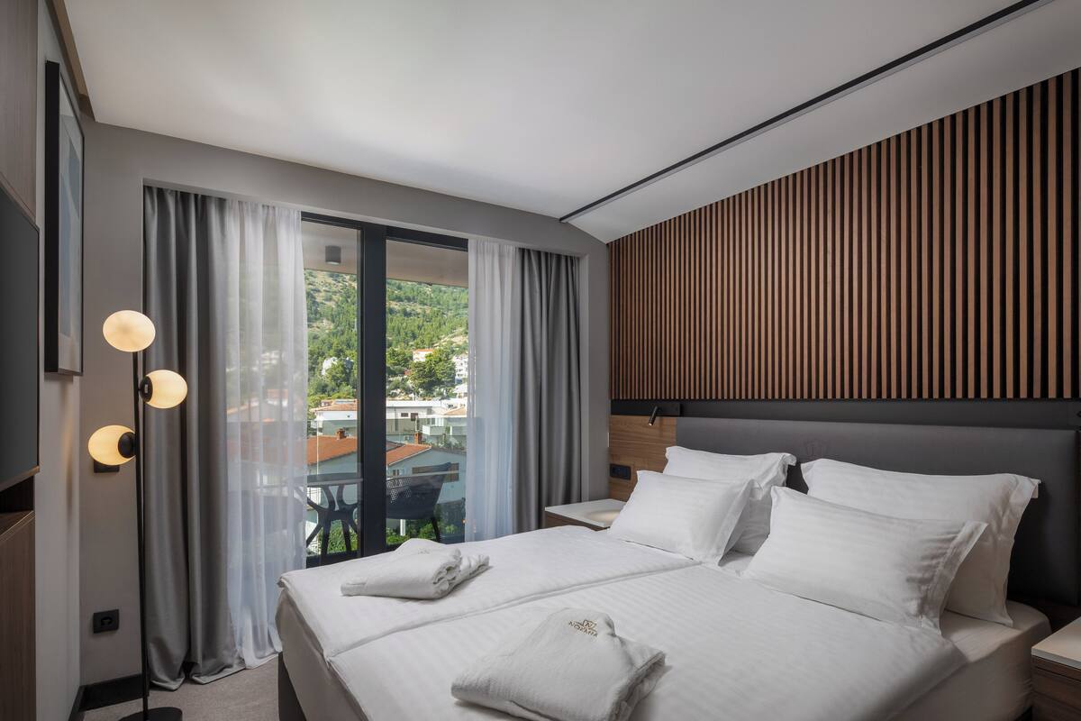 A modern hotel room with a large bed, a wooden headboard, and a view of the city through floor-to-ceiling windows.