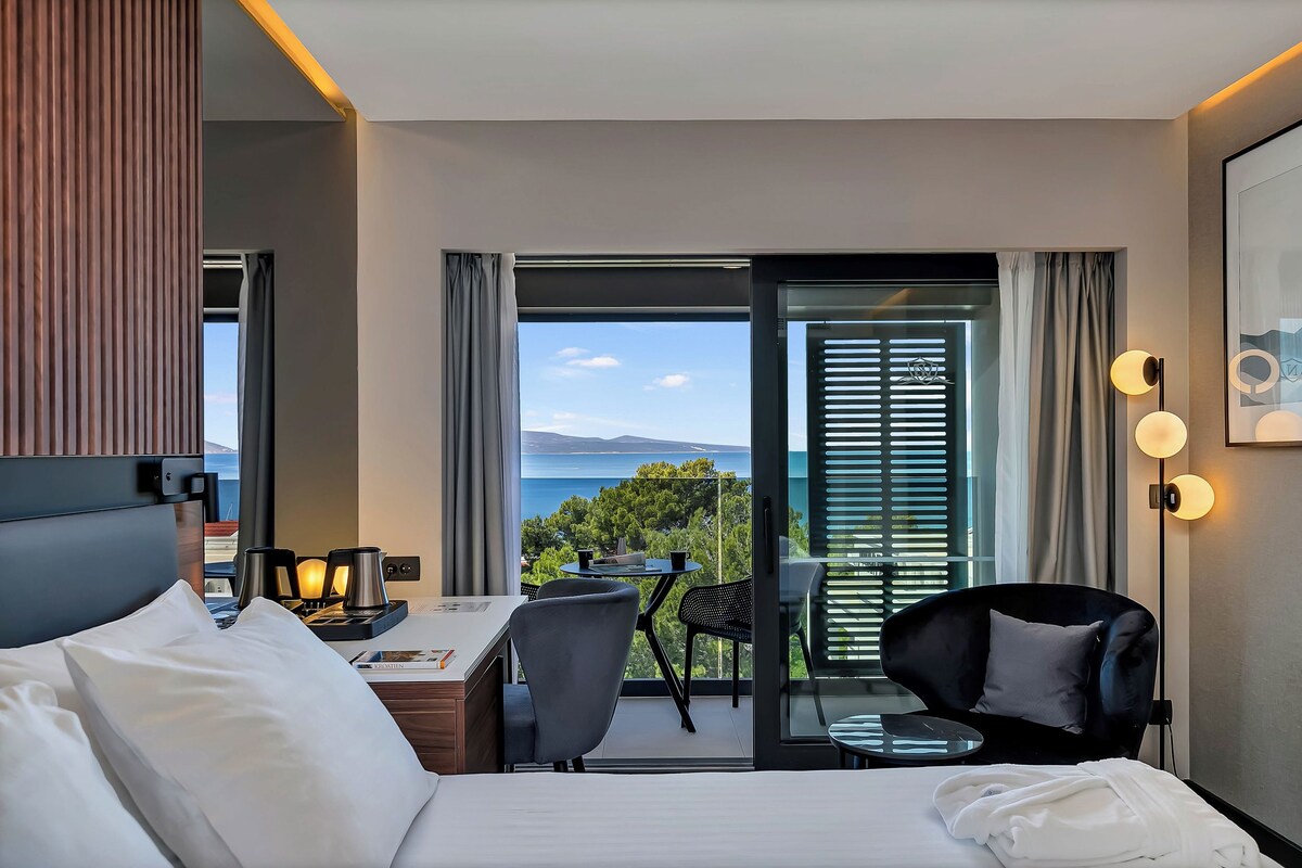 A hotel room with a bed, a desk, a chair, a small table, and a balcony with a view.