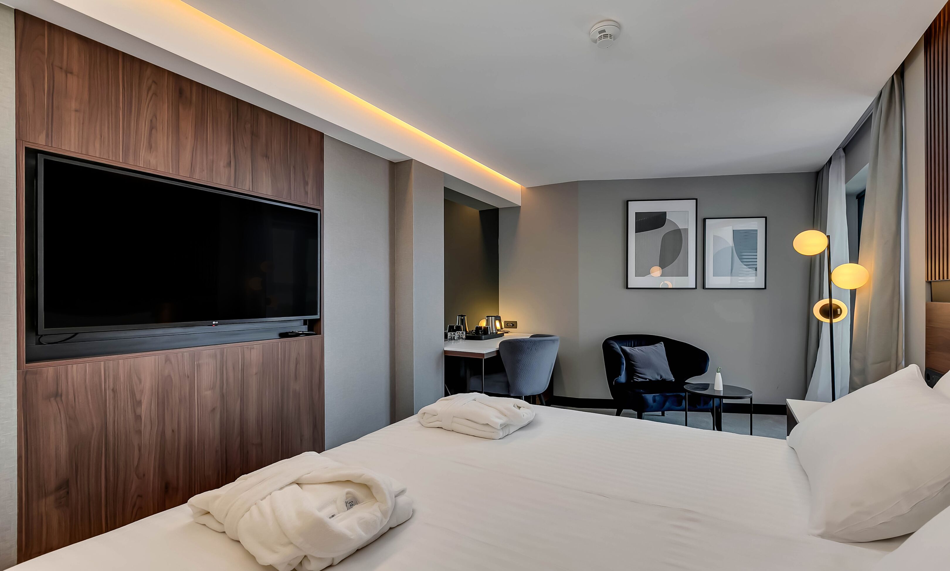 Ein Hotelzimmer mit großem Flachbildfernseher, einem Schreibtisch mit Stuhl, einem Bett mit Kissen und einem Nachttisch.