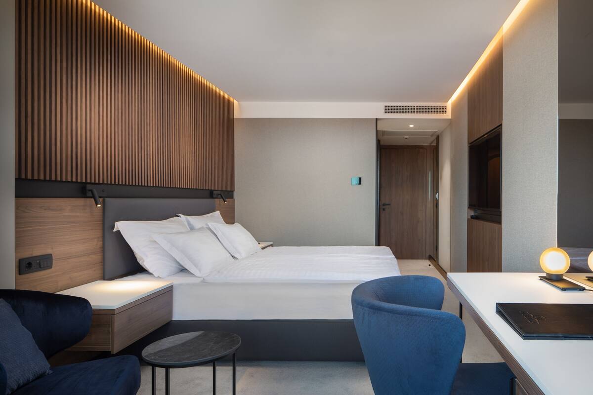 A modern hotel room with a bed, bedside table, chair, desk, and a TV.