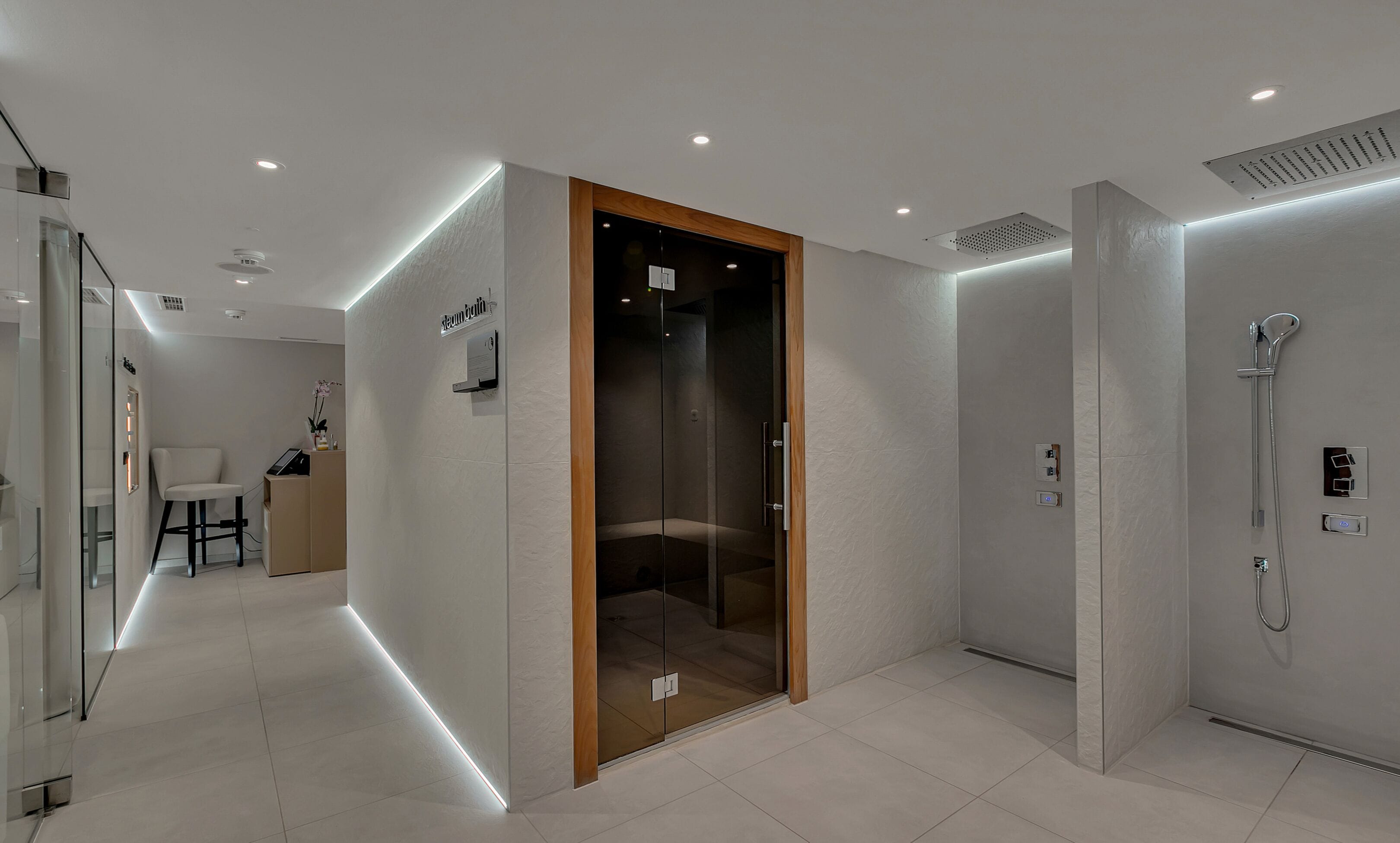 Un baño moderno con ducha de vidrio, tocador con espejo y una zona de estar con una silla y una mesita.