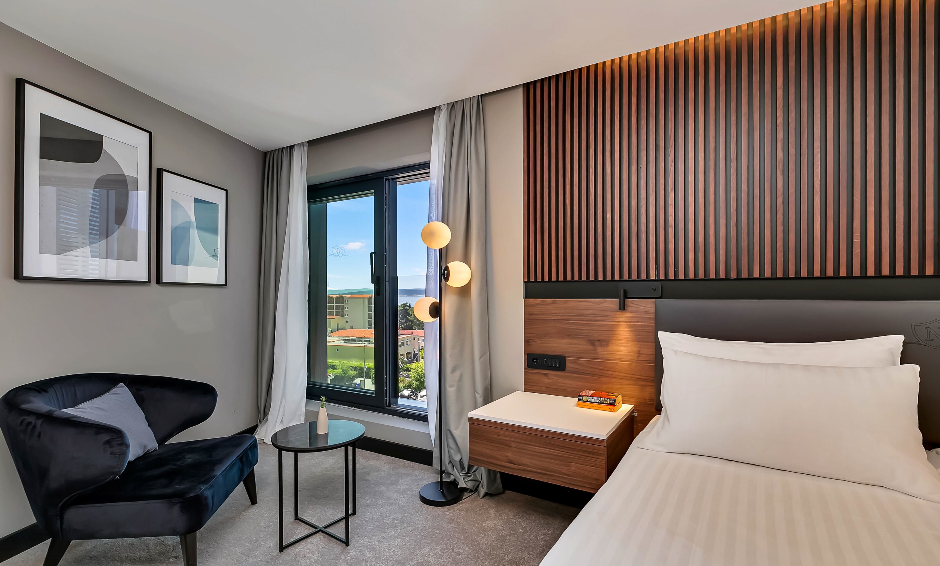 A hotel room with a bed, a desk, a chair, a small table, a window with curtains, and framed artwork on the wall.