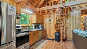 Une cuisine rustique aménagée dans une cabane en bois, avec un réfrigérateur, un four à micro-ondes et un four en acier inoxydable. On y trouve un escalier en bois et une fenêtre donnant sur un paysage verdoyant.