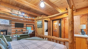 L’intérieur d’une cabane en rondins, avec un lit, des fauteuils verts, une cheminée et un téléviseur.