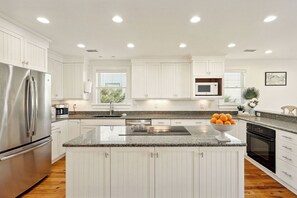 A modern kitchen with white cabinetry, stainless steel appliances, granite countertops, and a central island with a bowl of fruit. - East Arctic Avenue 0603 - Sunshine By The Sea (Folly Beach)