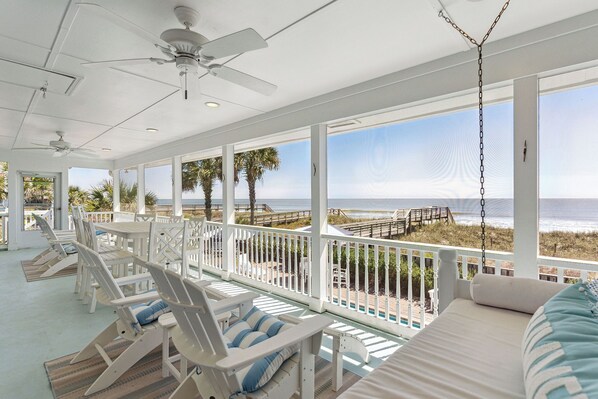 A covered outdoor seating area with white chairs and a sofa, overlooking a beach with a wooden walkway.