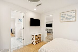 Ein Schlafzimmer mit einem Bett, einem an der Wand befestigten Fernseher, einer weißen Kommode und einer Tür zu einem anderen Raum.