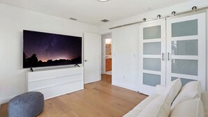 A modern living room with a flat-screen TV, a grey ottoman, a white sofa, and a sliding glass door closet. - Sunset Dock Pet-Friendly & Steps to Beach (Naples)