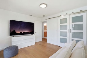 A modern living room with a flat-screen TV, a grey ottoman, a white sofa, and a sliding glass door closet.