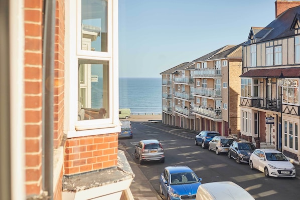 A coastal residential street with parked cars, a view of the sea, and multi-story buildings with balconies.