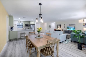 A modern kitchen with a dining area, a living room with a sofa and TV, and a large window.