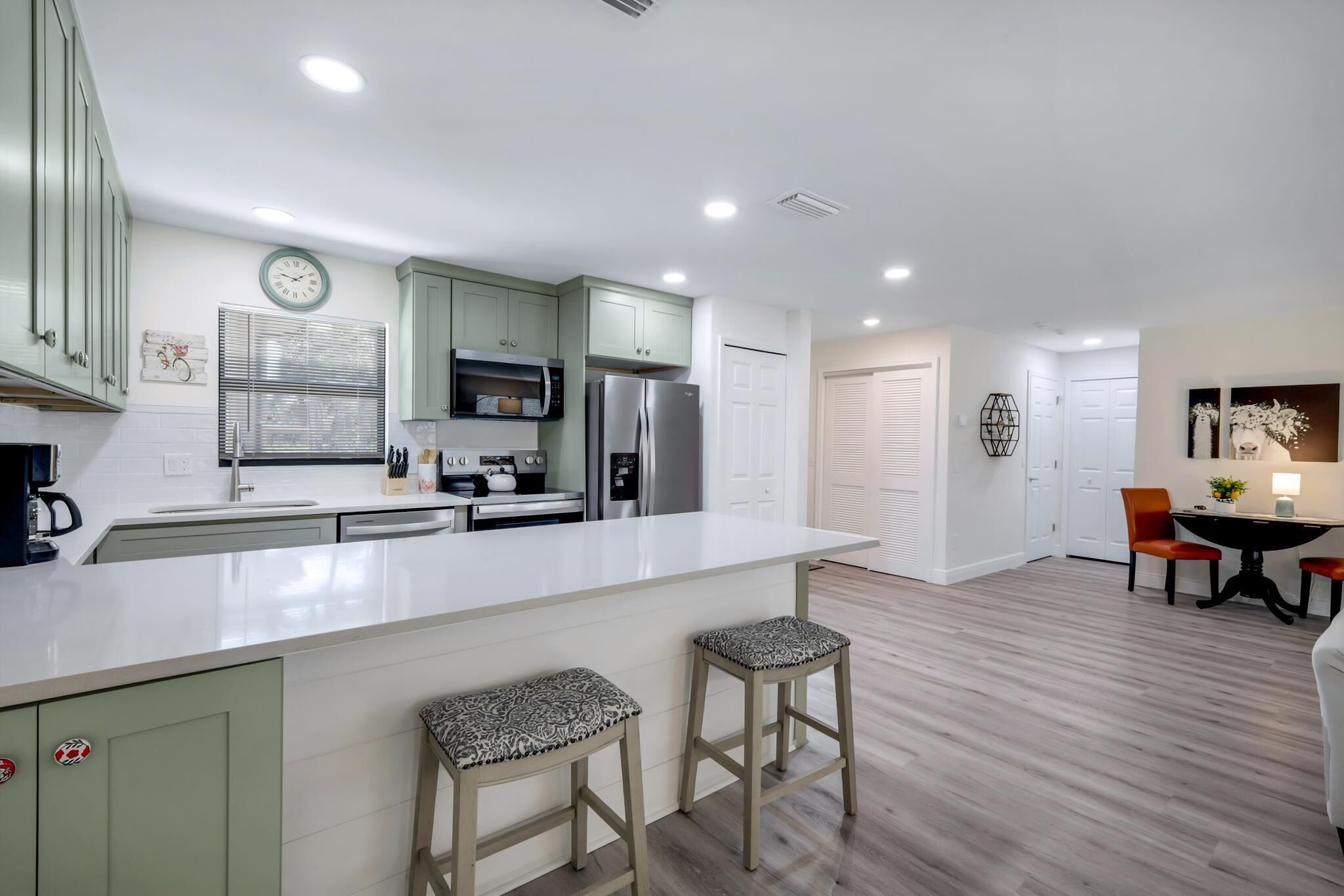 A modern kitchen with a white countertop, green cabinets, and a stainless steel refrigerator. There are two stools and a dining table with chairs.