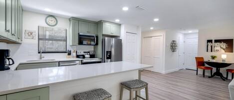 A modern kitchen with a white countertop, green cabinets, and a stainless steel refrigerator. There are two stools and a dining table with chairs.