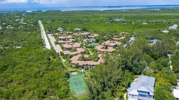 An aerial view of a residential area with houses arranged in a heart shape, surrounded by dense greenery and a tennis court.