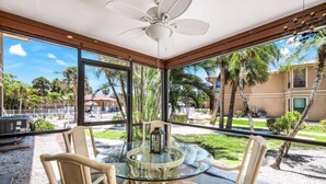 Une terrasse fermée par des baies vitrées, avec une table ronde et des chaises, un ventilateur de plafond et une vue sur les palmiers et les immeubles.