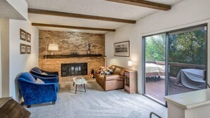 Un salon avec une cheminée en briques, des fauteuils bleus, un canapé marron et une terrasse avec des meubles d’extérieur.
